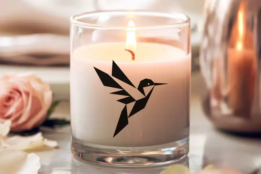 A lit candle in a clear glass jar, featuring a geometric hummingbird design.