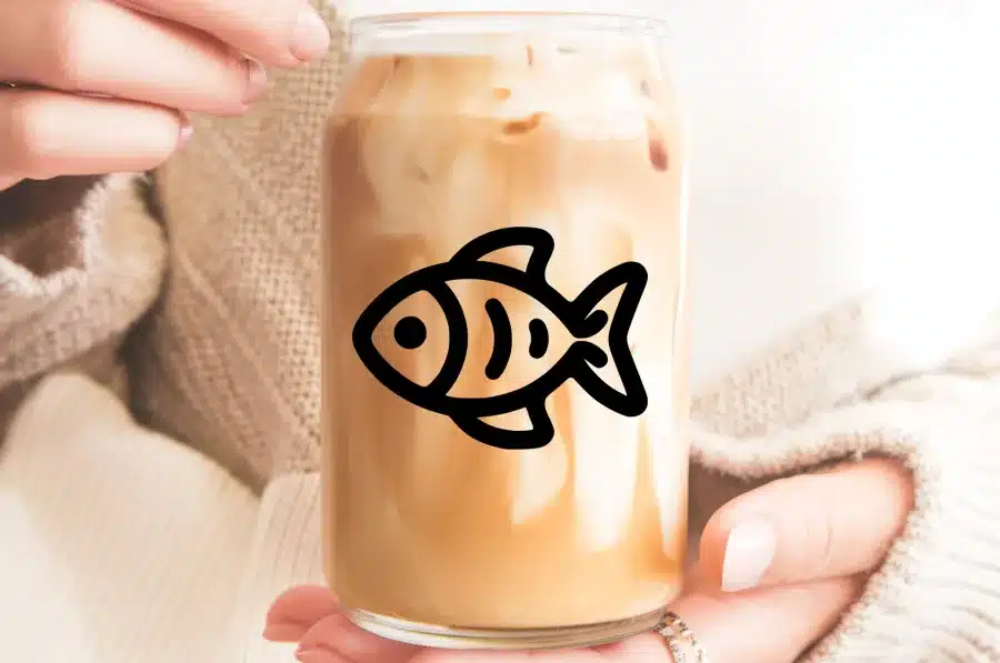 A Person'S Hands Holding A Glass Iced Coffee With A Fish Design On It.