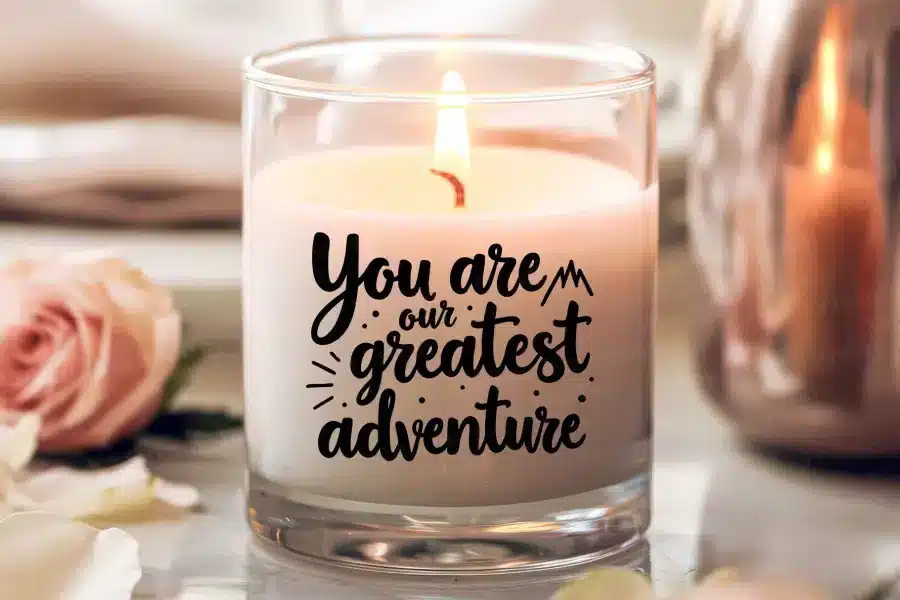 A Lit Candle In A Clear Glass Jar With The Text You Are Our Greatest Adventure.