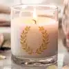 A Lit Candle In A Clear Glass Jar With A Gold Laurel Wreath Design.