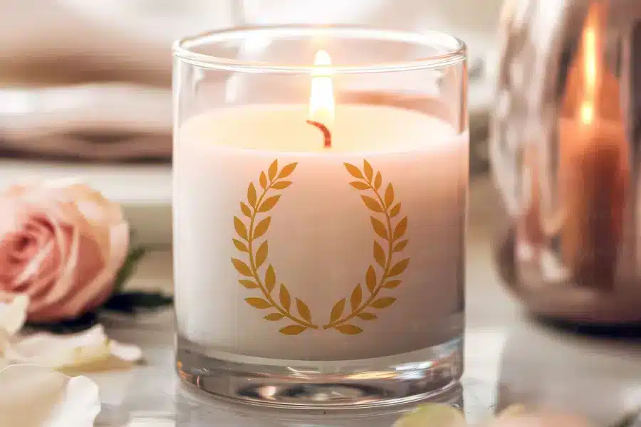 A lit candle in a clear glass jar with a gold laurel wreath design.