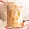 A Person'S Hands Hold A Glass Iced Coffee Can With A Gold Laurel Wreath Design.