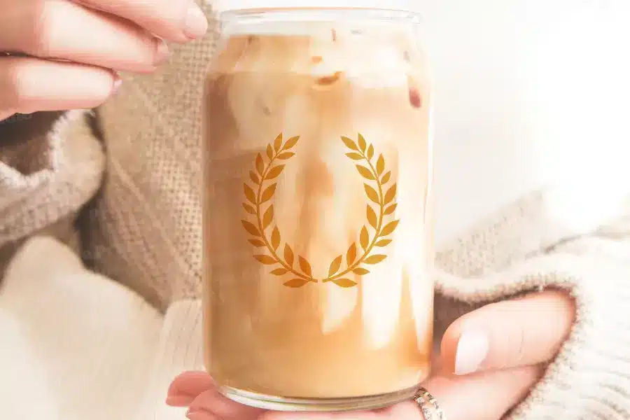 A Person'S Hands Hold A Glass Iced Coffee Can With A Gold Laurel Wreath Design.