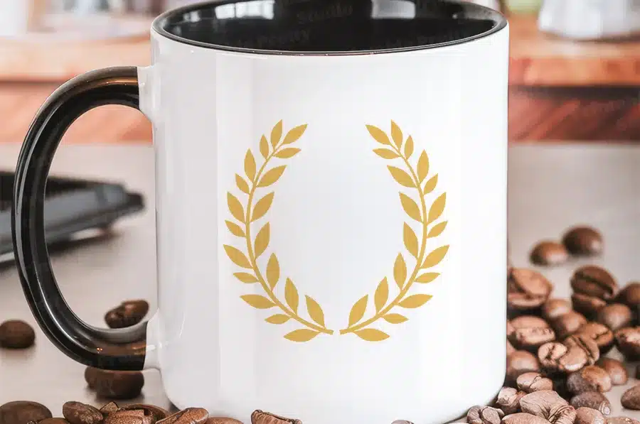 White Coffee Mug With Black Handle, Featuring A Gold Laurel Wreath Design.