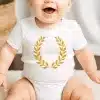 Baby Wearing A White Onesie With A Gold Laurel Wreath Design.