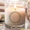 A Lit Candle In A Clear Glass Jar With A Green Leaf Wreath Design.