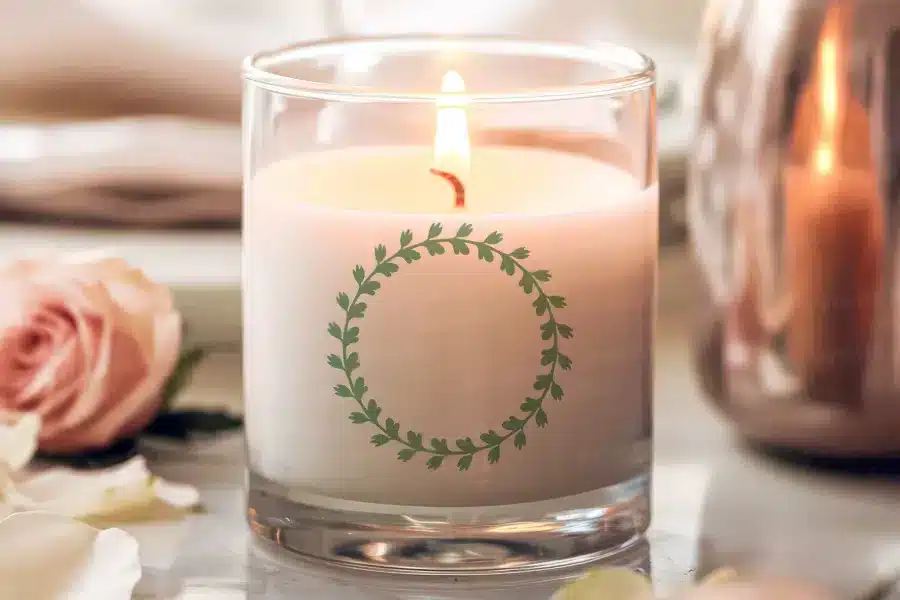 A lit candle in a clear glass jar with a green leaf wreath design.