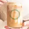 A Person'S Hands Hold A Glass Iced Coffee Can With A Green Leafy Wreath Design.