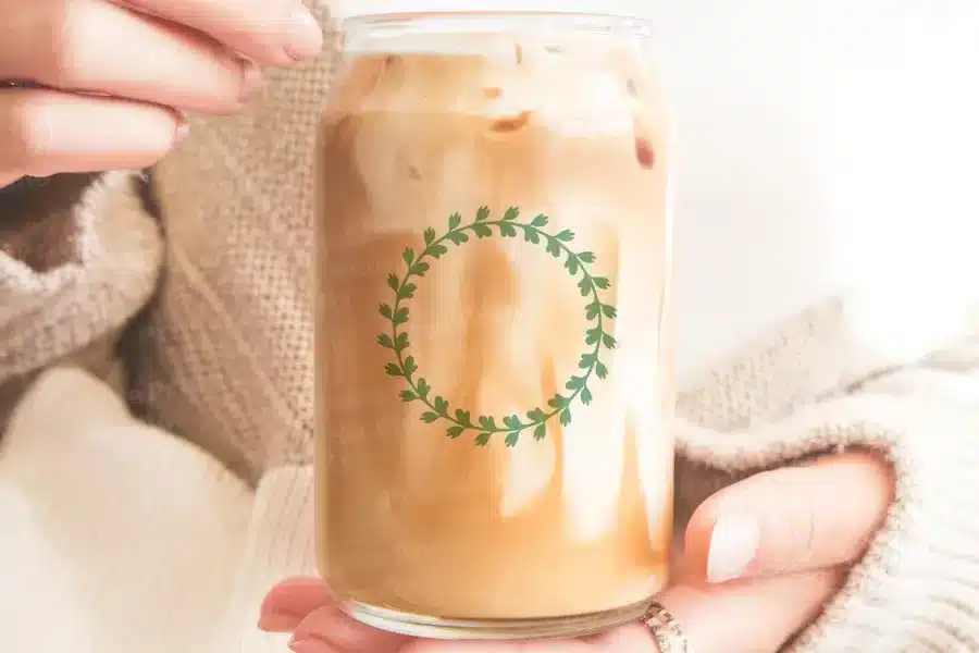 A Person'S Hands Hold A Glass Iced Coffee Can With A Green Leafy Wreath Design.