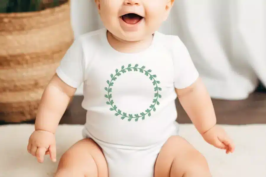 Baby Wearing A White Bodysuit With A Green Leafy Wreath Design.