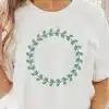 Woman Holding Iced Coffee, Wearing A White T-Shirt With A Sage Green Leafy Wreath Design.