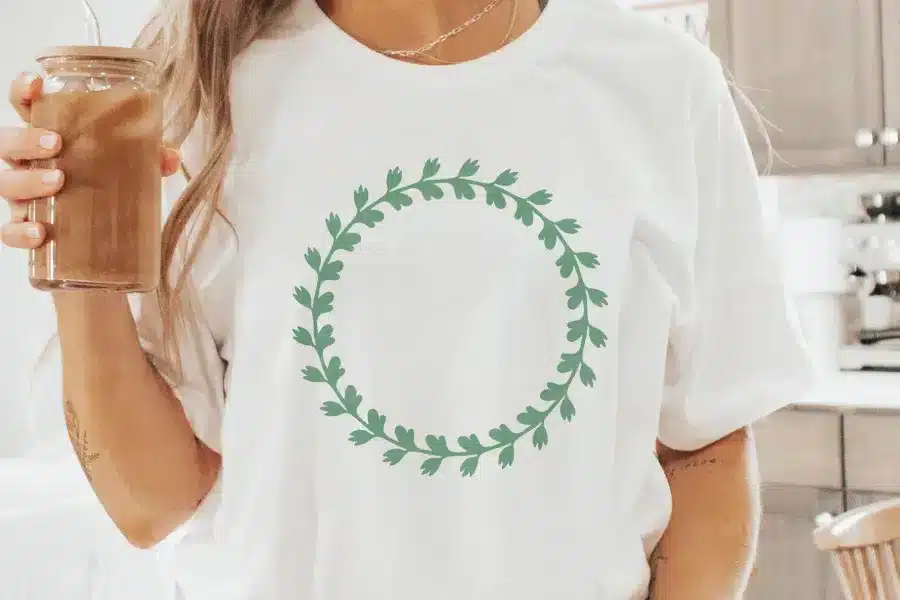 Woman Holding Iced Coffee, Wearing A White T-Shirt With A Sage Green Leafy Wreath Design.