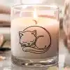 A Lit Candle In A Glass Jar Features A Line Drawing Of A Sleeping Cat.