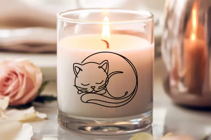 A Lit Candle In A Glass Jar Features A Line Drawing Of A Sleeping Cat.
