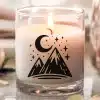 A Lit Candle In A Clear Glass Jar Features A Design Of Mountains, A Crescent Moon, And Stars.
