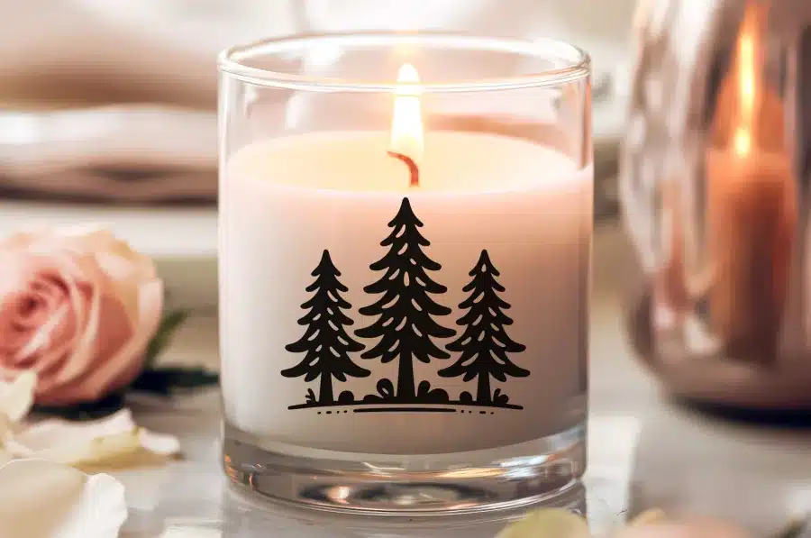 A Lit Candle In A Glass Jar Decorated With Three Black Evergreen Trees.