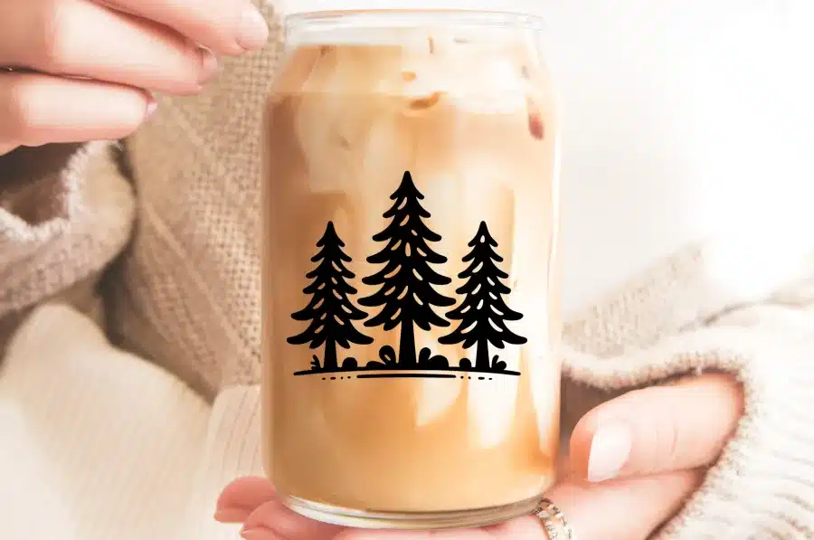 Hands Holding A Glass Of Iced Coffee With A Design Of Three Pine Trees.