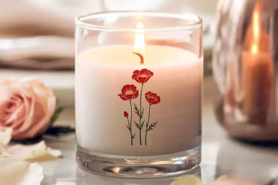 A lit candle in a clear glass jar, decorated with a simple design of three red poppies.