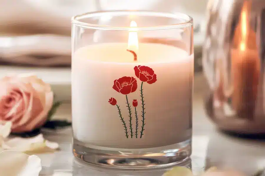 A lit candle in a clear glass jar with a poppy flower design.