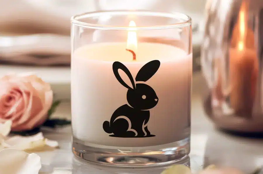 A Lit Candle In A Glass Jar Features A Black Bunny Rabbit Design.