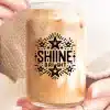 A Person Holds A Glass Jar Filled With Iced Coffee. The Jar Features The Words Shine Bright In A Decorative Font With Stars.