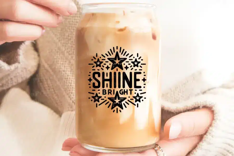 A Person Holds A Glass Jar Filled With Iced Coffee. The Jar Features The Words Shine Bright In A Decorative Font With Stars.