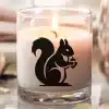 A Lit Candle In A Glass Jar Features A Black Silhouette Of A Squirrel Holding A Nut.