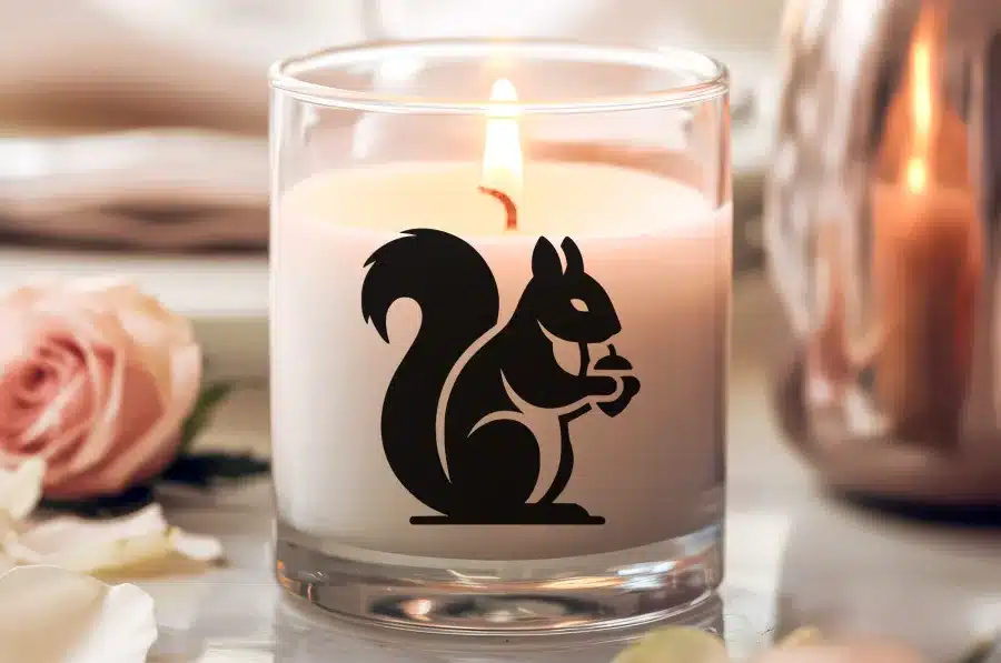 A Lit Candle In A Glass Jar Features A Black Silhouette Of A Squirrel Holding A Nut.