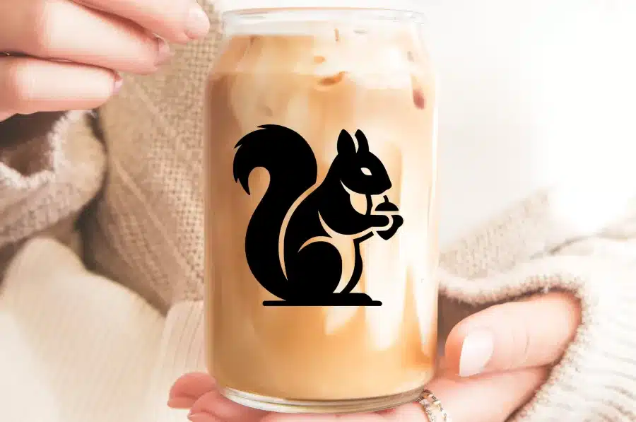 A Person'S Hands Holding An Iced Coffee In A Glass Jar With A Black Silhouette Of A Squirrel Holding An Acorn.