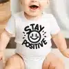 Baby Wearing A White Onesie With Black Text That Says Stay Positive And A Smiley Face.