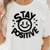 Woman Wearing A White T-Shirt With A Black Design That Says Stay Positive And Includes A Smiley Face.