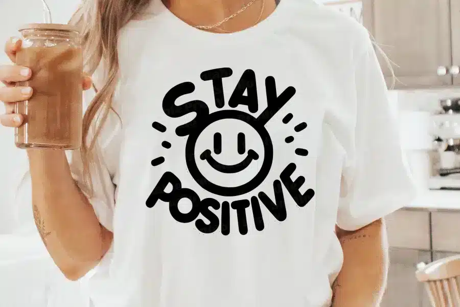 Woman Wearing A White T-Shirt With A Black Design That Says Stay Positive And Includes A Smiley Face.