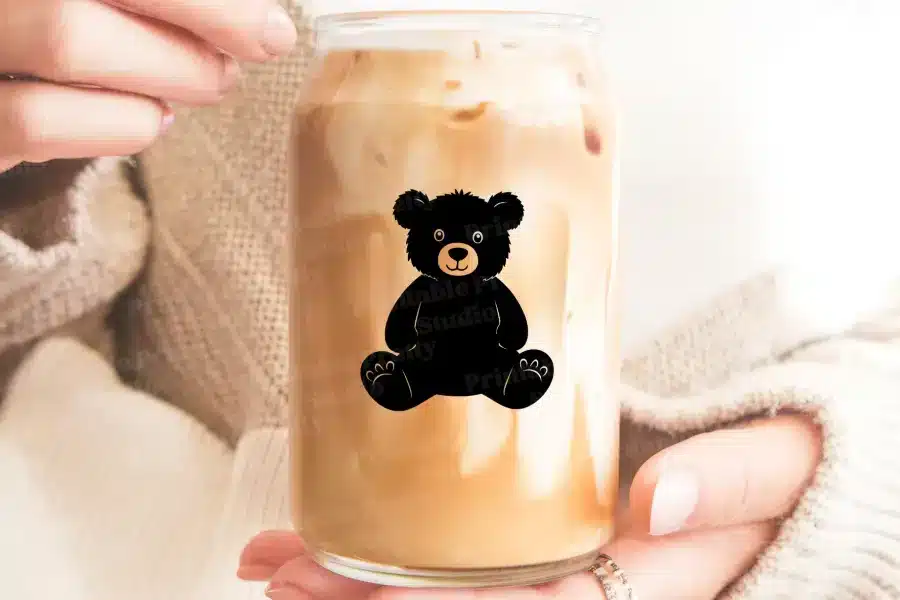 A Person Holds A Glass Iced Coffee With A Black Teddy Bear Design.