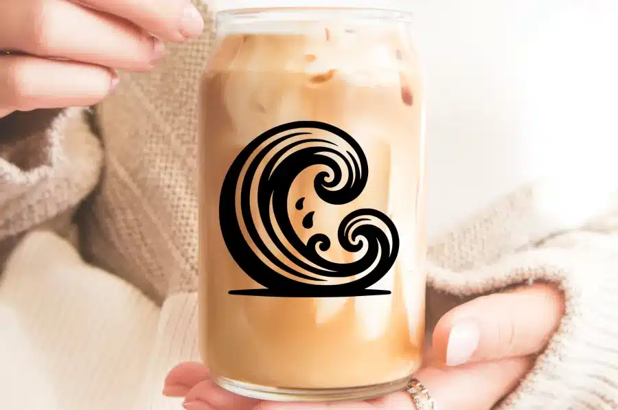 A Person Holds A Glass Jar Filled With Iced Coffee.  A Stylized Wave Design Is On The Jar.