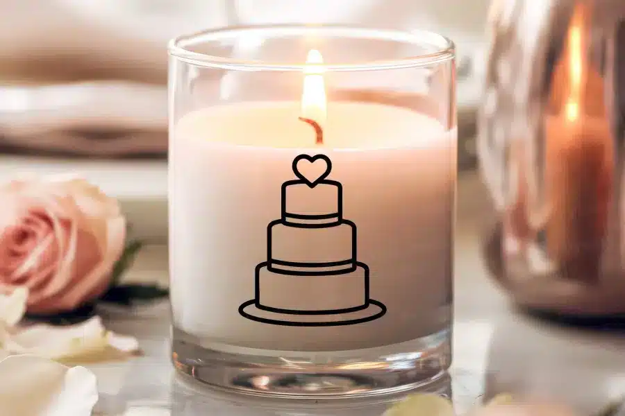 A lit candle in a clear glass jar, decorated with a simple line drawing of a three-tiered wedding cake topped with a heart.