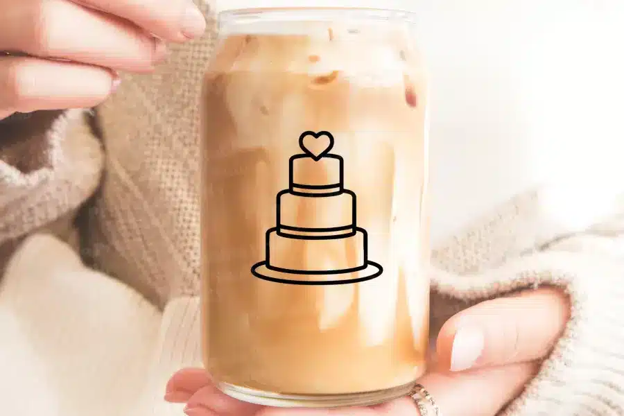 A Person Holds A Glass Jar Filled With Iced Coffee. A Simple Line Drawing Of A Wedding Cake With A Heart On Top Is On The Jar.