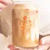 A Person'S Hands Hold A Glass Jar Filled With Iced Coffee. An Orange Floral Design Is On The Jar.