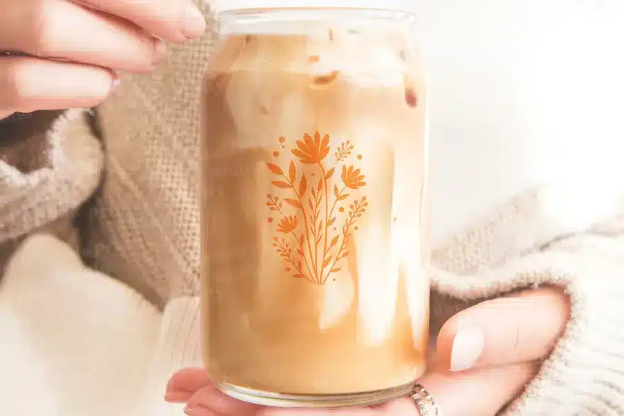A Person'S Hands Hold A Glass Jar Filled With Iced Coffee. An Orange Floral Design Is On The Jar.
