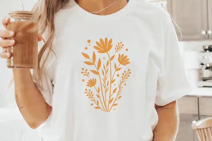 Woman holding iced coffee, wearing a white t-shirt with a mustard yellow floral design.